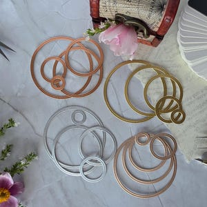 Puede incluir: Cuatro juegos de troqueles circulares de metal en acabados cobre, oro, plata y oro rosa. Cada juego contiene anillos anidados con un detalle de borde punteado, dispuestos sobre una superficie clara junto a unas tijeras y papel.