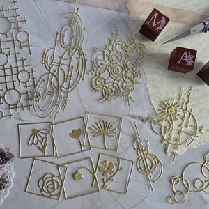 Könnte beinhalten: Eine Sammlung goldfarbener Metallstanzteile mit geometrischen Gittern, abstrakten kreisförmigen Mustern und quadratischen Rahmen mit Blumenmotiven. Drei kleine rote Holzblöcke mit den weißen Buchstaben M, A und E sind enthalten.
