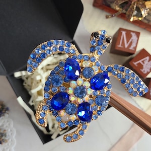 Könnte beinhalten: Eine schildkrötenförmige Brosche mit einem goldfarbenen Metallrahmen, der mit zahlreichen kleinen hellblauen Strasssteinen, größeren ovalen dunkelblauen Kristallen und einer zentralen Gruppe irisierender, facettierter Steine besetzt ist.