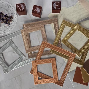 May include: A collection of square metal frames in silver, gold, and copper tones, featuring intricate etched border patterns. The set includes various sizes of open frames and solid square inserts, accompanied by three dark brown blocks with the letters M, A, and E.