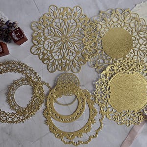 Könnte beinhalten: Eine Sammlung goldfarbener, runder Papierspitzendeckchen mit komplizierten Stanzmustern. Das Set enthält verschiedene Größen und Designs, einige mit festen Mitten und andere mit durchbrochenen Mustern, angeordnet neben kleinen Holzblöcken und einer Schere.