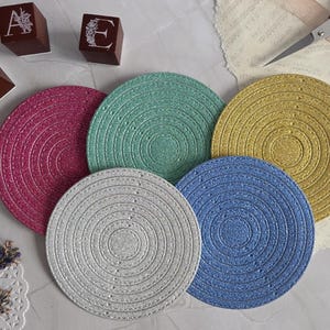 Puede incluir: Cinco posavasos circulares con un acabado texturizado y brillante en tonos magenta, verde menta, oro, plata y azul. Cada posavasos presenta un patrón de espiral concéntrica. Se observan tres bloques de madera marrón con las letras M, A y E.