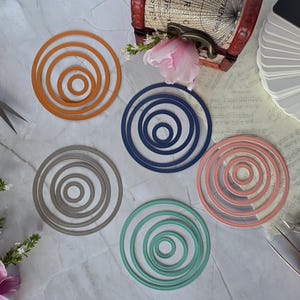 Puede incluir: Cinco juegos de troqueles de corte circulares de metal anidados en naranja, azul marino, gris, verde azulado y rosa polvoriento. Cada juego consta de varios anillos concéntricos con un patrón de borde punteado sobre una superficie.