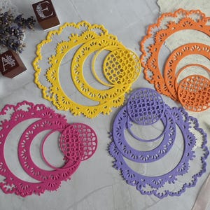 May include: Four sets of circular die-cut paper shapes in yellow, orange, pink, and purple. Each set contains nested rings with intricate lace-like scalloped edges and geometric grid patterns. Three small wooden blocks with letters E, A, and M are visible.