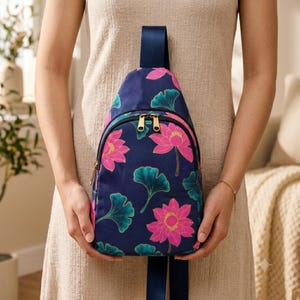 May include: A navy blue sling backpack featuring a vibrant pattern of bright pink lotus flowers and teal ginkgo leaves. The bag has a single dark blue shoulder strap, a front zippered pocket with gold-toned hardware, and a rounded top silhouette.