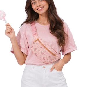 May include: A pink fanny pack featuring a delicate floral pattern with small red and white flowers, a gold-toned zipper closure, and an adjustable pink strap, worn across the chest over a plain pink t-shirt and white denim shorts.