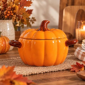 May include: A bright orange ceramic pumpkin-shaped lidded tureen with a glossy finish. The vessel features a curved brown stem handle on the lid and two brown side handles. The exterior is ribbed, mimicking the natural texture of a pumpkin.