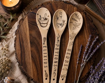 Könnte beinhalten: Drei helle Holzlöffel mit schwarzen, lasergravierten Illustrationen und Schriftzügen. Der erste Löffel zeigt einen Mörser mit Stößel und das Wort ELIXIRS. Der zweite zeigt eine Flasche mit POTIONS. Der dritte zeigt Kräuter mit dem Wort HERBS.