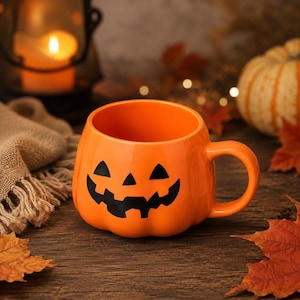 Könnte beinhalten: Ein leuchtend orangefarbener Keramikbecher in Kürbisform mit einem schwarzen Jack-o'-Lantern-Gesicht, bestehend aus dreieckigen Augen und einem gezackten Mund. Der Becher hat eine glatte, glänzende Oberfläche und einen geschwungenen Henkel an der rechten Seite.