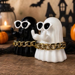 Peut inclure: Deux figurines de fantômes, une noire et une blanche, portant chacune des lunettes de soleil à monture blanche et une chaîne dorée autour du milieu. Les deux personnages ont les bras levés avec les doigts tendus, finition lisse et brillante.