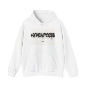 Puede incluir: Una sudadera blanca de manga larga con capucha que presenta un gráfico rectangular central con el texto HYPERFOCUS en negro estilo grafiti y el texto más pequeño ESTD MM debajo. La prenda incluye un bolsillo tipo canguro y capucha.