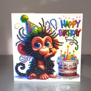 May include: A square greeting card featuring a diamond art monkey with large blue eyes, wearing a colourful party hat. The monkey sits next to a tiered birthday cake with lit candles. The text HAPPY BIRTHDAY is displayed in colourful gems.
