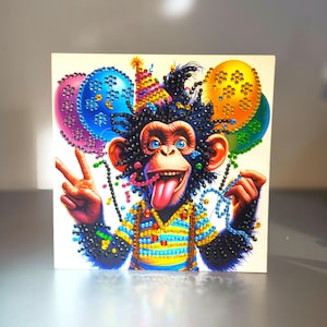 May include: A square diamond painting kit featuring a chimpanzee with a party hat, tongue out, and a peace sign gesture. The image is decorated with colourful balloons in blue, pink, yellow, and green, accented with sparkling rhinestones throughout.