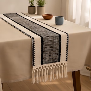Puede incluir: Un camino de mesa de color crema con una franja central negra texturizada con patrón tejido, bordeado por un fino ribete geométrico negro y terminado con largos flecos anudados de color crema en los extremos de la tela.