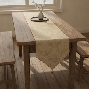 May include: A rectangular beige table runner with a subtle, repeating wave-like pattern and a pointed end featuring a single hanging tassel. The runner is centred on a wooden table flanked by two wooden benches, with a small vase holding white flowers.