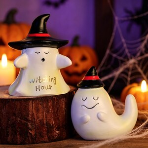May include: Two white ghost figurines with closed eyes and black witch hats featuring red bands. The larger ghost has the text "witching Hour" printed in black on its front. Both figures have smooth, rounded shapes and are crafted from ceramic.