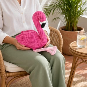 May include: A bright pink crocheted flamingo plush toy featuring a long, curved neck, a black and white beak, and light pink legs. The stuffed animal is crafted with a textured yarn stitch pattern and has closed eyes embroidered in black thread.