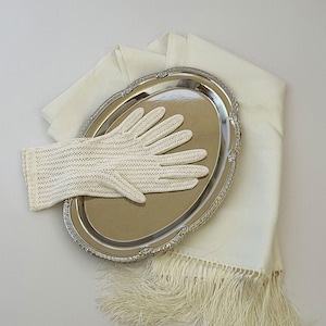 May include: A pair of off-white crochet gloves with vertical ribbed detailing resting on a polished silver-tone oval tray, which is placed atop a folded off-white fabric shawl featuring long, delicate fringe along the bottom edge.