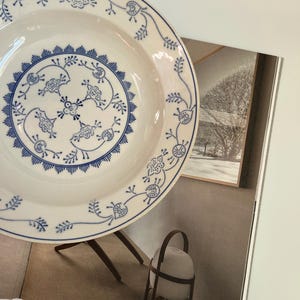 May include: A white ceramic plate featuring a blue floral pattern. The design includes a central circular motif with stylised flowers and leaves, surrounded by a decorative border of radiating lines and a vine pattern along the outer rim.