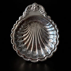 May include: A polished silver-toned dish shaped like a scallop shell. The bowl features deep, radiating fluted grooves and a scalloped edge. The top section is decorated with an ornate, embossed scrollwork pattern with a central floral motif.