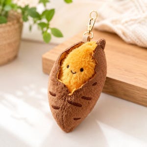 May include: A plush keychain shaped like a brown sweet potato with a split skin revealing a bright yellow, fuzzy interior. The yellow center features a small embroidered face with two black eyes and a curved mouth. A gold metal lobster clasp is attached.