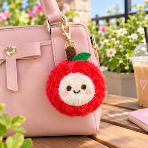 May include: A light pink handbag with a gold heart-shaped accent and a fuzzy, round, red apple-shaped keychain. The keychain features a white face with dark eyes, a small smile, pink cheeks, a brown stem, and a green leaf, attached by a gold clasp.