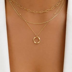 May include: A layered gold-tone necklace set consisting of three distinct chains. The top chain features elongated oval links, the middle is a fine curb chain, and the bottom chain holds a small, circular, twisted metal pendant charm.