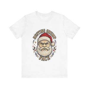 May include: A white short-sleeved t-shirt featuring a graphic of a grumpy Santa Claus wearing a red hat. The text RESTING GRUMP FACE is arched around the illustration, which is framed by small holly leaves and stars in red, green, and gold.