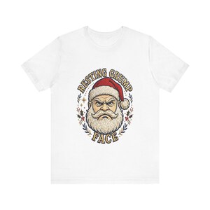 Könnte beinhalten: Ein weißes Kurzarm-T-Shirt mit einer Grafik eines mürrischen Weihnachtsmanns mit roter Mütze. Der Text RESTING GRUMP FACE ist um die Illustration gebogen, die von kleinen Stechpalmenblättern und Sternen in Rot, Grün und Gold umrahmt wird.