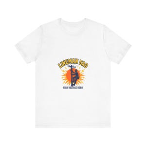 May include: A white short-sleeved t-shirt featuring a graphic print of a person climbing a utility pole against an orange sunburst background. The text reads LINEMAN DAD in yellow arched letters and HIGH VOLTAGE HERO in blue text below.