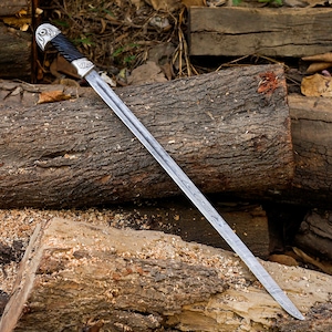 May include: A shashka sword featuring a long, slightly curved steel blade with a visible fuller. The hilt has a black, spirally grooved grip, a silver-colored pommel with an engraved bird motif, and a matching silver-colored crossguard ferrule.