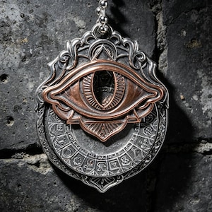 Puede incluir: Un colgante de metal plateado con un motivo de ojo central de color cobre sobre un disco circular grabado con varios símbolos. El colgante tiene un borde ornamentado de estilo filigrana y cuelga de una cadena de bolas de color plata.