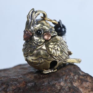 Peut inclure: Un pendentif figurine de calopsitte en laiton avec une finition dorée métallique, présentant une crête détaillée, des yeux sombres et des taches sur les joues de couleur cuivre. L'oiseau a une petite ouverture circulaire à la base.