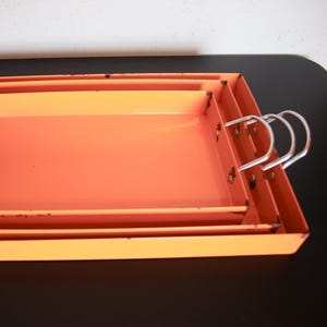 Op de afbeelding: Een set van drie in elkaar passende rechthoekige metalen dienbladen met een feloranje afwerking. Elk dienblad heeft opstaande randen en twee zilverkleurige metalen handgrepen die met klinknagels aan de zijkanten zijn bevestigd.