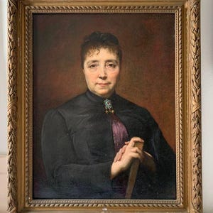 Può includere: Un ritratto a olio di una persona con capelli scuri che indossa un indumento nero a collo alto con un accento viola e una spilla decorativa. Il soggetto tiene in mano un oggetto di legno. Il dipinto è racchiuso in una cornice dorata intagliata.