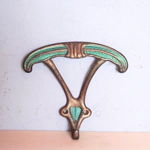 Può includere: Un gancio da parete con una barra superiore curva e un gancio centrale rivolto verso il basso. La superficie metallica presenta una finitura in bronzo anticato con vivaci accenti verde acqua che mettono in risalto i motivi a foglia sulla parte superiore e sulla base.