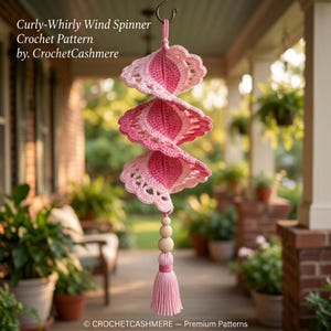 Puede incluir: Un patrón de ganchillo para molinillo de viento Curly-Whirly de CrochetCashmere. La decoración colgante presenta un diseño en espiral en tonos rosa y rosa claro, terminado con tres cuentas de madera y una borla de hilo rosa claro.