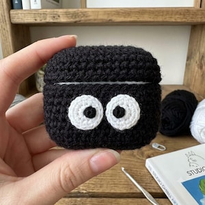 May include: A black crochet case for wireless earbuds featuring two large, circular white eyes with black pupils centered on the front. The textured, handmade cover is designed to fit snugly over the charging case, providing a soft, fuzzy exterior.