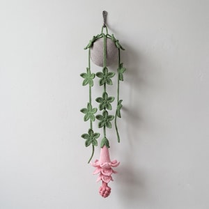 May include: A crocheted hanging plant featuring a light grey spherical base with long green vines adorned with five-petaled green flowers. The plant terminates in a layered, light pink bell-shaped blossom with a textured, rounded pink tip.