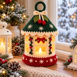 May include: A crocheted lantern featuring a dark green roof with white snowflake embroidery, a cream-colored body with red bow and berry accents, and a red base with white snowflake details. A small LED candle glows inside the structure.