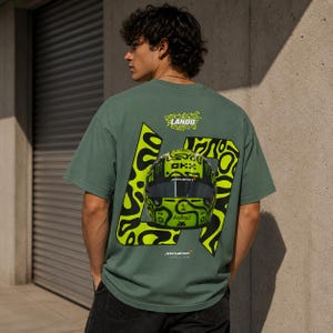 May include: A sage green short-sleeve t-shirt featuring a large graphic on the back. The design includes a bright neon yellow racing helmet with black patterns, the number 4, the text Lando, McLaren, and Android, all set against a black abstract shape.