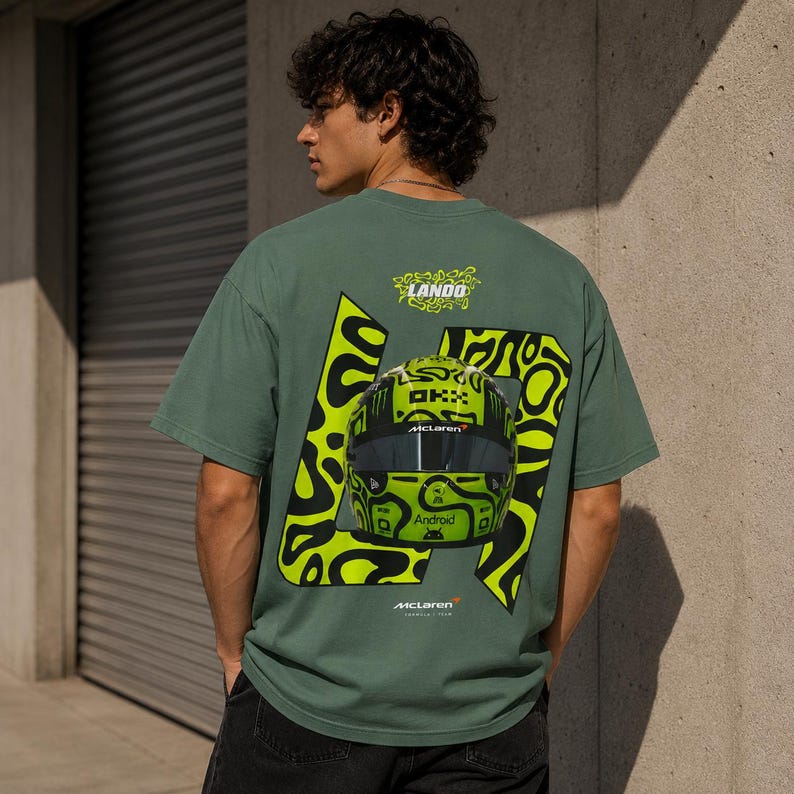 May include: A sage green short-sleeve t-shirt featuring a large graphic on the back. The design includes a bright neon yellow racing helmet with black patterns, the number 4, the text Lando, McLaren, and Android, all set against a black abstract shape.