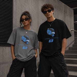 Può includere: Una t-shirt grigio antracite e una t-shirt nera, entrambe caratterizzate da una grafica di un casco da corsa blu, il testo "GEORGE RUSSELL" e il numero "63" in blu. Entrambe le magliette hanno maniche corte e girocollo, abbinate a pantaloni cargo neri.