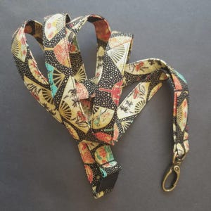 May include: A fabric lanyard featuring a black background with a repeating pattern of colorful folding fans in shades of red, yellow, teal, and cream. The strap includes a metallic bronze-toned swivel lobster clasp attached to one end.