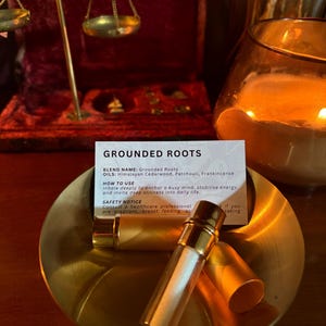 May include: A small gold-toned metal bowl containing two gold-capped glass essential oil vials and a white informational card labeled Grounded Roots. The card lists ingredients including Himalayan Cedarwood, Patchouli, and Frankincense.
