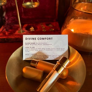May include: A gold-toned metal bowl containing two small cylindrical containers with gold caps and a white rectangular card. The card features the text DIVINE COMFORT, lists ingredients as Bergamot, Neroli, and Sandalwood, and includes usage instructions and a safety notice.