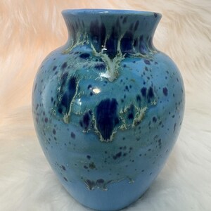 May include: A ceramic vase with a glossy finish, featuring a light blue base color with darker blue and deep indigo mottled spots and streaks that drip down from the rim, creating a textured appearance across the rounded body of the vessel.