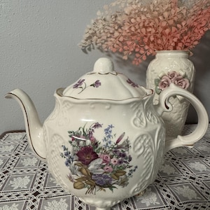 Könnte beinhalten: Eine cremefarbene Keramik-Teekanne mit einer bunten Blumenstrauß-Illustration auf der Vorderseite, geprägten Gittermustern an den Seiten, einem passenden Deckel, einem geschwungenen Griff, einem Ausguss und kleinen Füßen mit Goldrand.