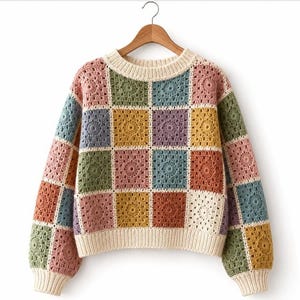Puede incluir: Un suéter de manga larga confeccionado con cuadrados de ganchillo multicolores en tonos rosa, verde, azul, amarillo y morado. La prenda presenta un cuello redondo, puños y dobladillo acanalados de color crema, colgado en una percha de madera.