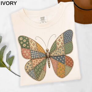 May include: An ivory t-shirt featuring a large butterfly graphic composed of various patchwork-style fabric patterns in shades of green, blue, yellow, and red. The shirt has a crew neckline and a Comfort Colors brand label at the inner neck.